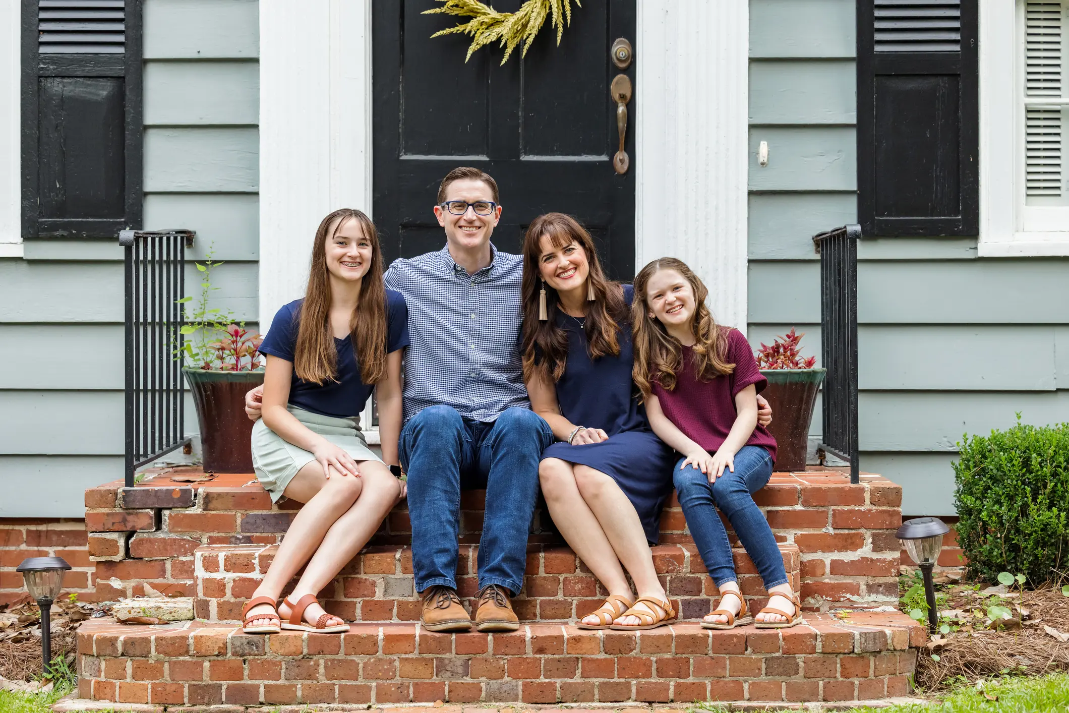 A family with a mother, father, and two daughters sitting outside on the brick porch of a small blue cottage house.
